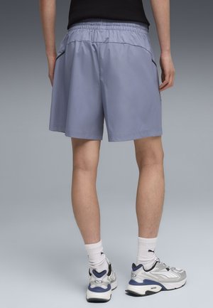 Mężczyzna ubrany w jasnoniebieskie sportowe spodenki, białe skarpetki i szare buty do biegania, stojący tyłem na szarym tle.