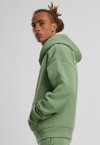 Haut à capuche vert en matériau doux, avec une capuche à cordon de serrage, des manches longues, une poche avant et des poignets côtelés. Branding minimal sur le côté.
