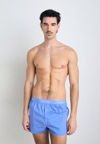 Modre bombažne boksarice z elastičnim pasom, ki ima bel logotip. Model stoji v nevtralnem položaju proti enobarvni ozadju.