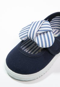 Zapatilla de lona navy con suela de goma blanca, que presenta un lazo a rayas azules y blancas en la punta y un forro interior a rayas.