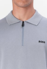 Világosszürke pólóing rövid cipzárral, puha kötött textúrával és fekete "BOSS" logóval a nyak bal oldala alatt.
