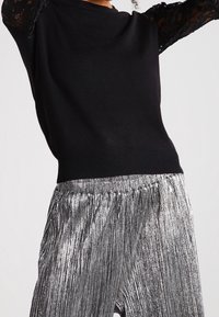 Osoba v černém svetru s krajkovými rukávy a stříbrných metalických plisovaných kalhotách na jednobarevném pozadí.