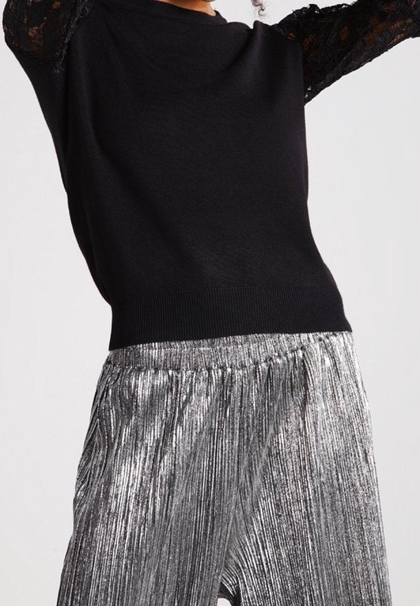 Osoba v černém svetru s krajkovými rukávy a stříbrných metalických plisovaných kalhotách na jednobarevném pozadí.