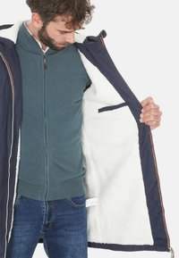 Giacca navy con un morbido rivestimento bianco, dotata di chiusura con zip e tasche laterali. Indossata sopra un maglione teal e jeans scuri.