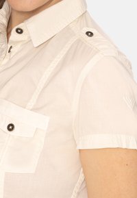 Camicia corta beige chiaro a maniche corte con colletto a bottoni, dotata di una tasca sul petto e bottoni scuri a contrasto, che presenta una texture liscia.