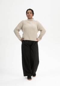 Helles beiges, geripptes Sweatshirt mit hohem Kragen, kombiniert mit schwarzen, weit geschnittenen Hosen und schwarzen Schuhen. Das Model steht selbstbewusst mit den Händen in den Hüften.