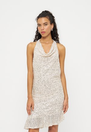 Femme aux cheveux bouclés portant une robe sans manches en sequins argentés avec un col bénitier et un ourlet à volants, debout devant un fond clair uni.
