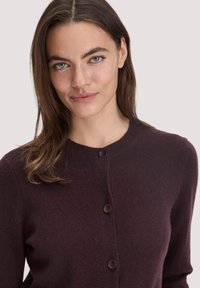 Burgundy cardigan valmistettu pehmeästä materiaalista, jossa on pyöreä kaula-aukko ja viisi nappia edessä sulkemiseksi. Suuri tekstuuri ja istuva muotoilu.