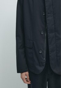 Marineblå letvægtsjakke med knapper foran, to forlommer og en lynlåslomme indvendigt. Glat stof med subtil struktur.