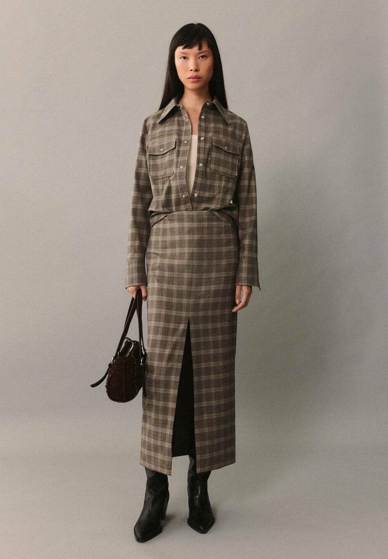 Ruskea ruudullinen pitkä hame, jossa on etublossi, yhdistettynä yhteensopivaan napitettavaan paitaan. Varustettu pitkillä hihoilla ja kahdella rintataskulla. Mustat nilkkurit ja pieni laukku viimeistelevät asun.