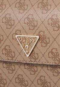 Hnedá textúrovaná taška cez rameno s opakujúcim sa vzorom, na prednej strane s zlatou logom doskou v tvare trojuholníka označenou "GUESS USA".