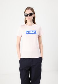 Lys rosa bomulls t-skjorte med en blå rektangulær "HUGO" logo. Modellen har på seg svarte kattøy solbriller og marineblå bukser. Enkel design med rund hals.