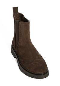 Bottines en suède marron avec des panneaux latéraux élastiques, des détails en brogue et une semelle en caoutchouc noir texturé. Équipées d'une languette à l'arrière.