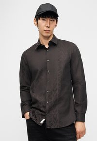 Νέος άνδρας που φοράει μαύρο πουκάμισο με σχέδια και μαύρο καπέλο, στέκεται με το ένα χέρι στην τσέπη μπροστά σε απλό φόντο.