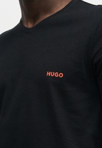 Μαύρο t-shirt με στρογγυλή λαιμόκοψη και μικρό φωτεινό πορτοκαλί λογότυπο "HUGO" στο αριστερό στήθος που φοριέται από άτομο με σκουρόχρωμο δέρμα.