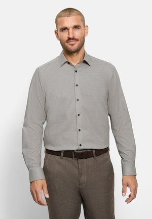 Mann trägt ein hellgraues, langärmeliges Hemd mit Knöpfen und braune Hose mit passendem braunen Gürtel, steht vor einem einfarbigen Hintergrund.