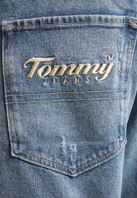 Ljusblå jeans i denim med en bakficka som har en broderad "Tommy Jeans"-logotyp i beige. Sliten detalj ovanför fickan.