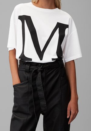 Personne portant un t-shirt blanc avec une grande lettre M noire et un pantalon noir taille haute avec ceinture nouée et grandes poches.