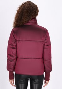 Burgundi pufi dzseki magas gallérral, strukturált steppelt mintával és bordázott mandzsettával. Simogatós, fényes anyagból készült, laza szabásban.