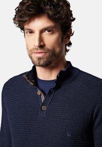 Granatowy sweter z dzianiny z teksturowanym wzorem, z placketem na pół guzika i kontrastowymi detalami kołnierza. Małe logo na piersi.