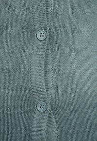 Pletený cardigan v jemné, světle šedé barvě s texturovaným povrchem. Obsahuje zapínání na přední straně se dvěma šedými knoflíky a žebrovanými okraji.