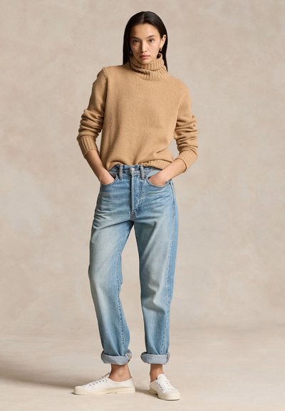 Nő bézs garbó pulóverben, világoskék felhajtott farmerral és fehér tornacipővel, zsebre tett kézzel áll, bézs háttér előtt.