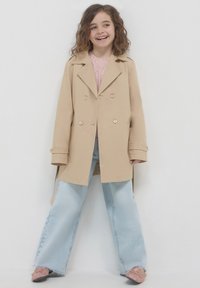 Beige Doppelreiher mit breiten Revers und goldenen Knöpfen, kombiniert mit hellblauen, weiten Jeans und rosa Ballerinas.