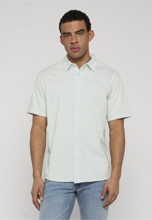 Homme aux cheveux courts et bouclés portant une chemise à manches courtes vert menthe clair boutonnée et un jean bleu clair, debout devant un fond uni.