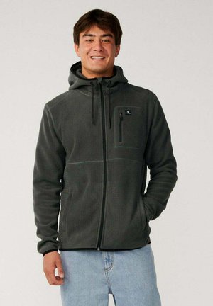Hombre que lleva una chaqueta de forro polar con cremallera, capucha y bolsillo en el pecho de color gris oscuro, sonriendo y vaqueros azul claro, de pie contra un fondo liso.