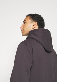 Ciemnofioletowy hoodie z zaokrąglonym kapturem i prążkowanymi mankietami. Miękka tekstura, widoczne detale szwu oraz luźny krój sylwetki.