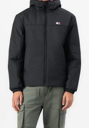 Mann trägt eine schwarze, wattierte Kapuzenjacke mit Frontreißverschluss und elastischen Bündchen, kombiniert mit olivgrünen Cargo-Hosen.