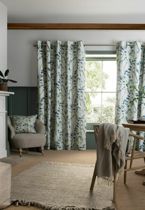 Vorhänge mit Blumenmuster bedecken ein Fenster, daneben befindet sich ein beiger Sessel mit passendem Kissen, ein Holzstuhl mit einer drapierten Decke und ein gewebter Teppich.
