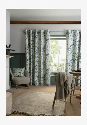 Vorhänge mit Blumenmuster bedecken ein Fenster, daneben befindet sich ein beiger Sessel mit passendem Kissen, ein Holzstuhl mit einer drapierten Decke und ein gewebter Teppich.
