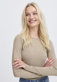 Blonde Frau mit langen Haaren in einem beigen Langarmshirt und blauen Jeans, lächelt und schaut zur Seite mit verschränkten Armen.