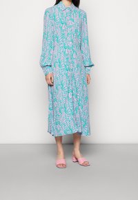 Robe chemise à manches longues de couleur lilas clair avec un motif ondulé turquoise et rose. Elle présente un devant boutonné et une coupe décontractée, accompagnée de mules roses.