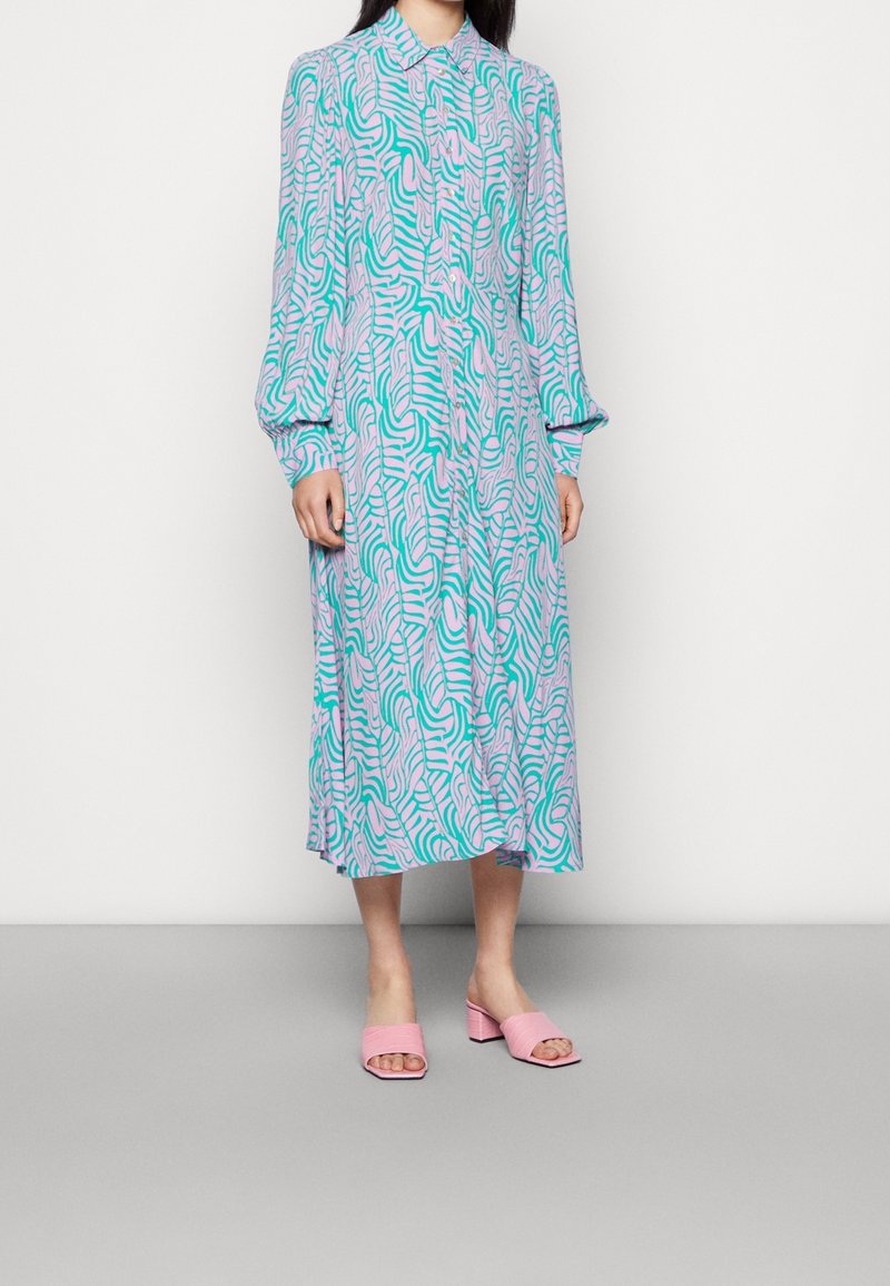 Robe chemise à manches longues de couleur lilas clair avec un motif ondulé turquoise et rose. Elle présente un devant boutonné et une coupe décontractée, accompagnée de mules roses.