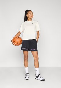 Αθλητική μπλούζα σε ανοιχτό μπεζ χρώμα με άνετη εφαρμογή, συνδυασμένη με μαύρο σορτς που έχει λουλουδάτο μοτίβο στο τελείωμα. Κρατάει μια μπάλα του μπάσκετ.