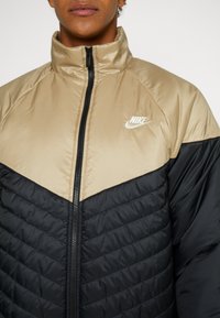 Chaqueta acolchada Nike con sección inferior negra y parte superior marr�ón claro, con cremallera frontal, cuello alto y costuras horizontales texturizadas.