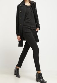 Manteau en laine noir avec boutons dorés sur un haut noir et un pantalon ajusté. Complété par des bottines noires, avec un bout et un talon pointus.