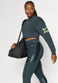 Mørkegrå cropped sweatshirt med Nike-logo; teal leggings med reflekterende accenter; model holder en sort træningstaske.
