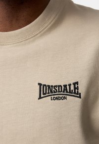 Bežové bavlnené tričko s čierne vyšitou logom "Lonsdale London" na hrudi, okrúhlym výstrihom a hladkou textúrou.
