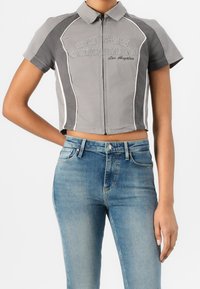 Harmaa lyhythihainen vetoketjullinen paita, jossa on kaulus ja valkoinen pipings. Edessä on brodeerattu "GUESS ORIGINALS Los Angeles".