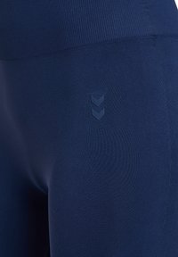 Close-up of dark blue stretchy fabric leggings with a small embossed double chevron logo on the upper thigh.