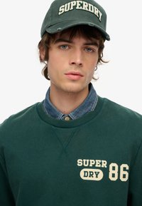 Zielona bluza z kapturem z napisem "SUPERDRY 86" w kolorze kremowym, zestawiona z dżinsową koszulą i pasującą zieloną czapką z logo "SUPERDRY".
