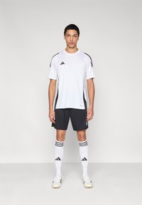 Bijela sportska majica s crnim naglascima, uparena s tamnim kratkim hlačama i bijelim čarapama s crnim prugama te crnim kopačkama.