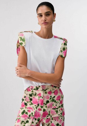 Donna con capelli scuri che indossa una maglia bianca con spalle imbottite a motivo floreale e pantaloni a vita alta abbinati con fiori rosa e verdi.