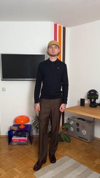 Άντρας με μπεζ καπέλο, μαύρο μπλουζάκι μακρύ μανίκι και καφέ παντελόνι που στέκεται σε ένα δωμάτιο με τηλεόραση, πορτοκαλί λάμπα και διακοσμημένο βαλιτσάκι.