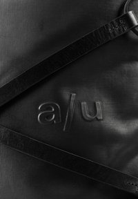 Schwarze, strukturierte Tasche mit zwei schwarzen Lederriemen und geprägten Buchstaben "a / u" auf der Oberfläche.