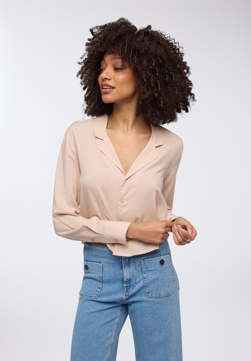 Blusa beige con bottoni e maniche lunghe, in tessuto morbido e design a V. Indossata con jeans azzurri a vita alta con tasche frontali.