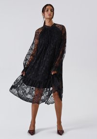 Abito di pizzo nero con maniche lunghe trasparenti, colletto alto, hem a volant e motivo floreale. Abbinato a décolleté bordeaux con punta affilata.